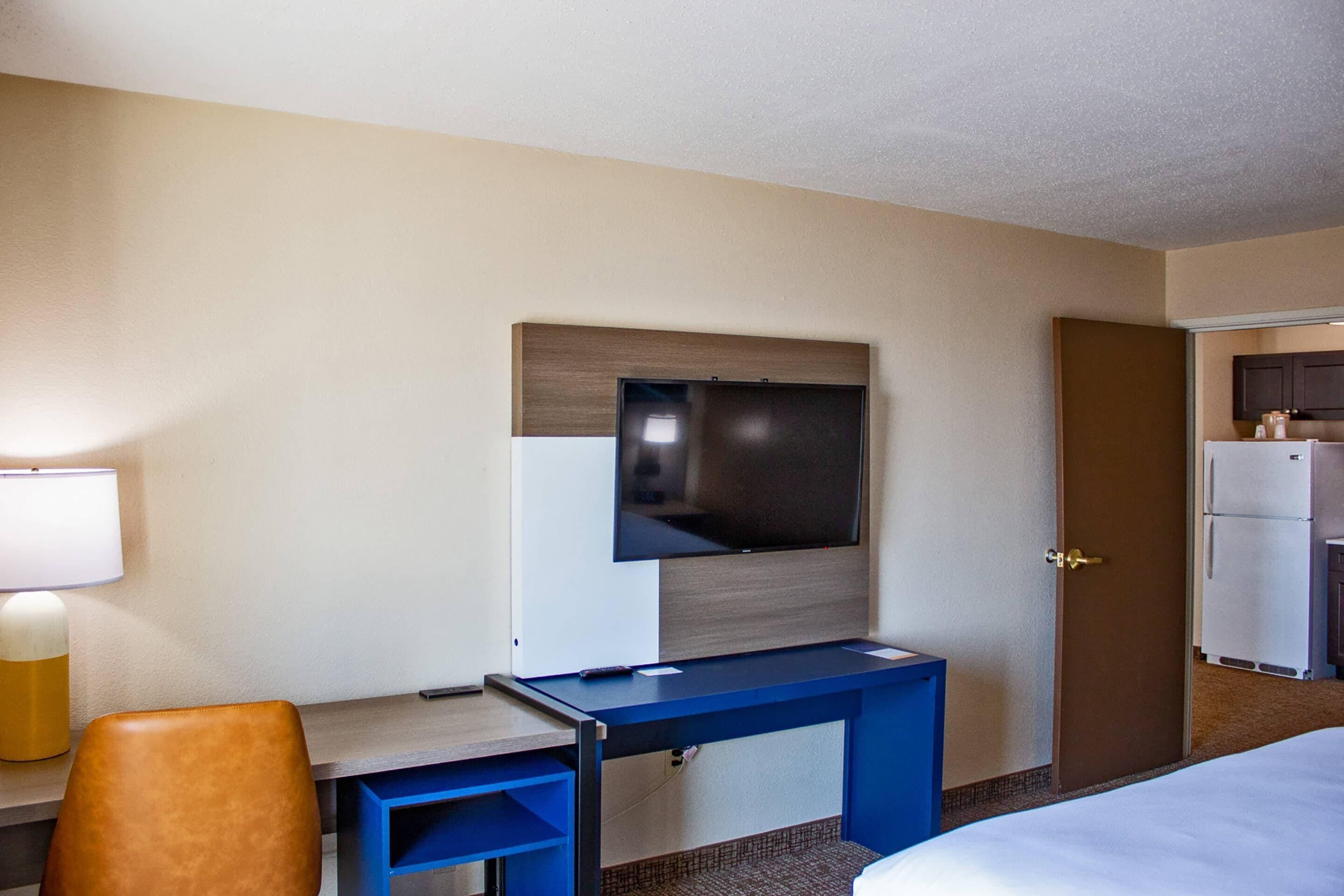 A hotel room with a bed, a desk with a chair, a television mounted on the wall, and a refrigerator.