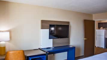 A hotel room with a bed, a desk with a chair, a television mounted on the wall, and a refrigerator.