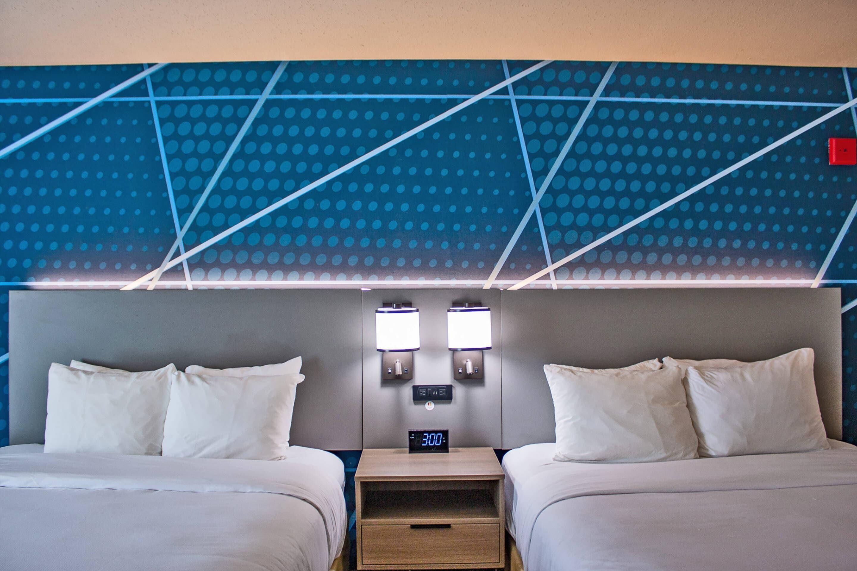 Habitación de hotel con dos camas, una mesita de noche y un reloj digital que muestra la hora.