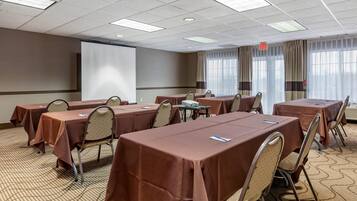 A conference room with multiple tables, chairs, a projector screen, and large windows with curtains.