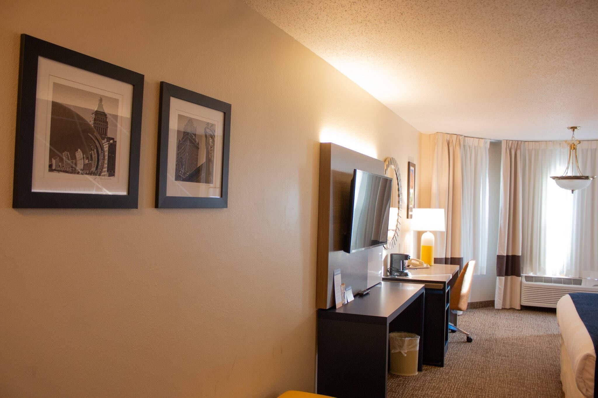 A hotel room with a bed, a desk with a computer, a TV, and two framed pictures on the wall.