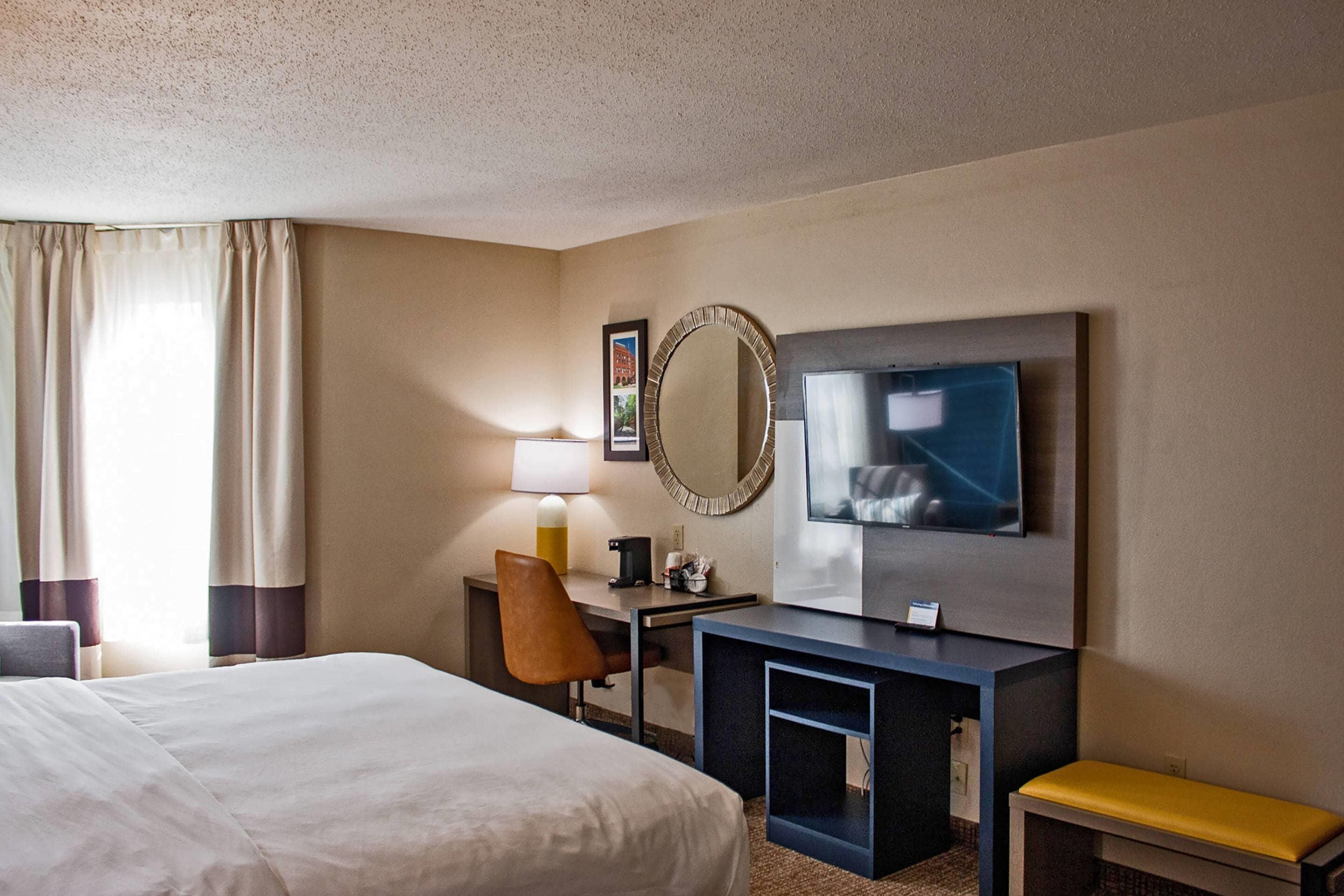 A hotel room with a large bed, a desk with a lamp, a television, and a mirror.