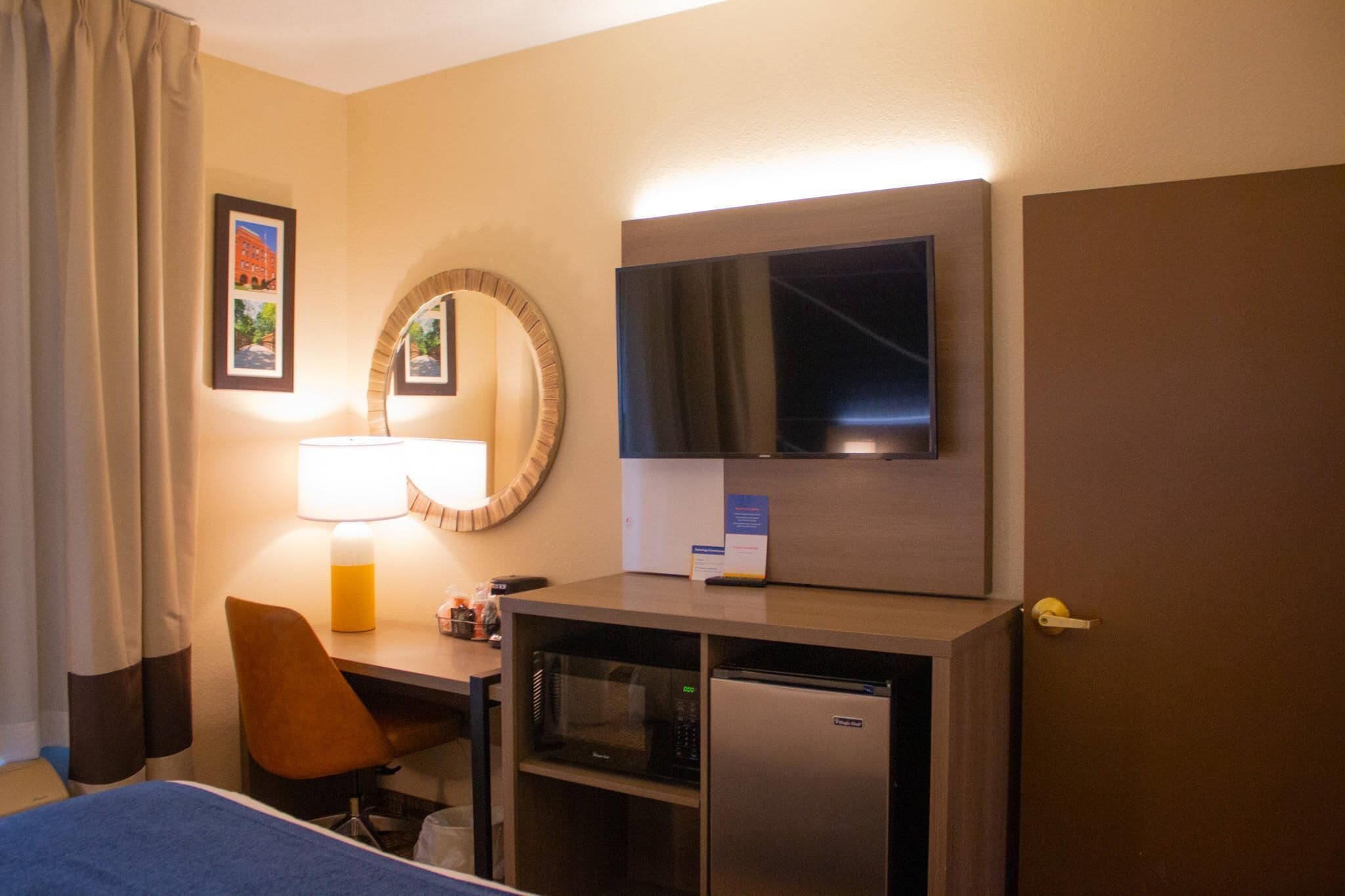 Habitación de hotel con cama, escritorio con lámpara, microondas, refrigerador, televisor y puerta.