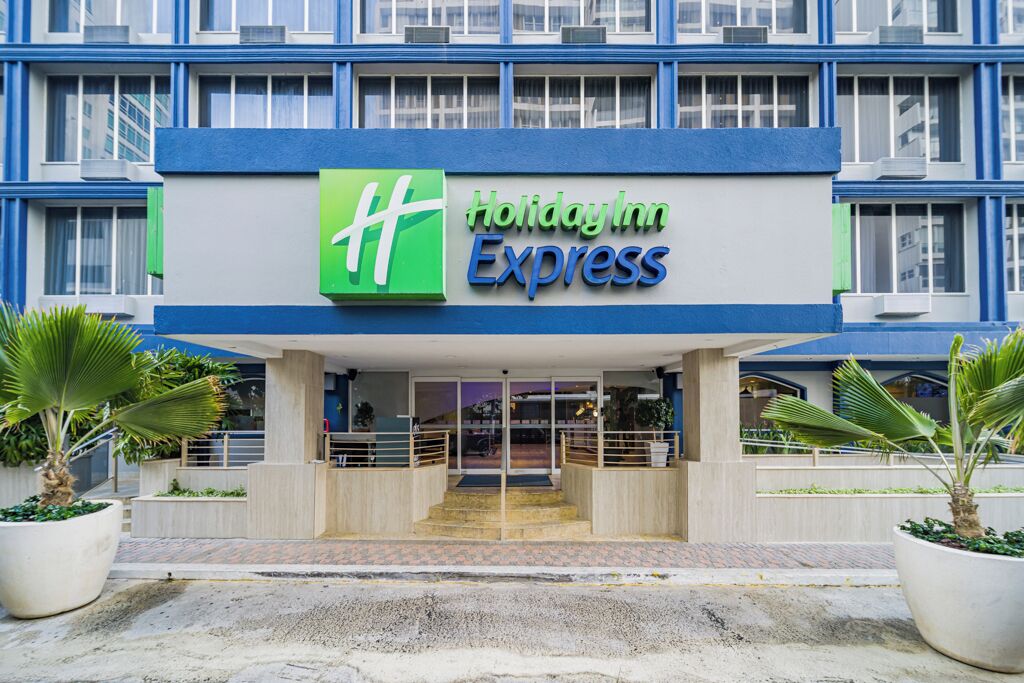 A Holiday Inn Express building with a prominent green and blue logo, a modern entrance with steps, and potted plants on either side.