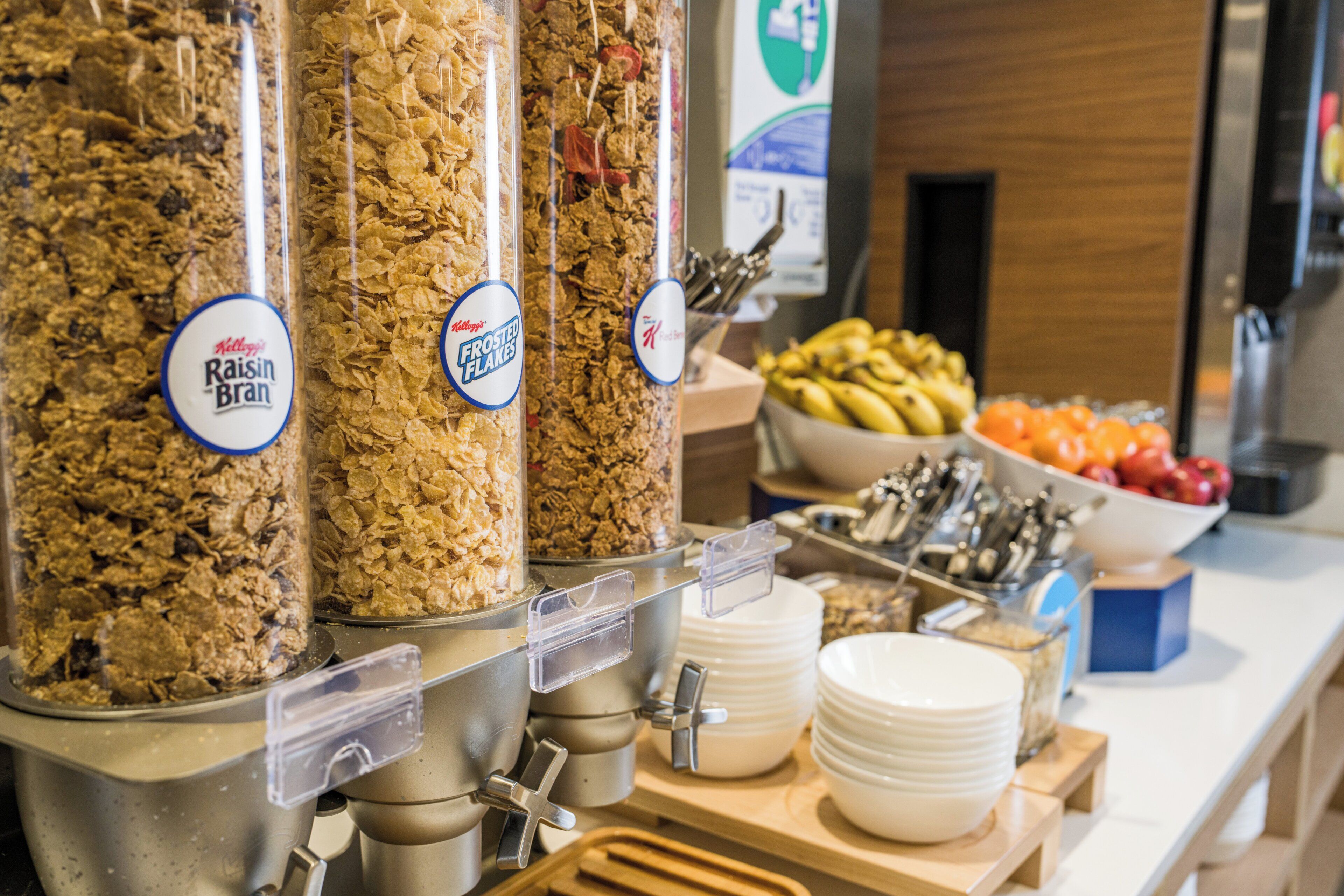 A breakfast buffet with cereal dispensers labeled 'Raisin Bran' and 'Frosted Flakes,' and a bowl of fruit in the background.