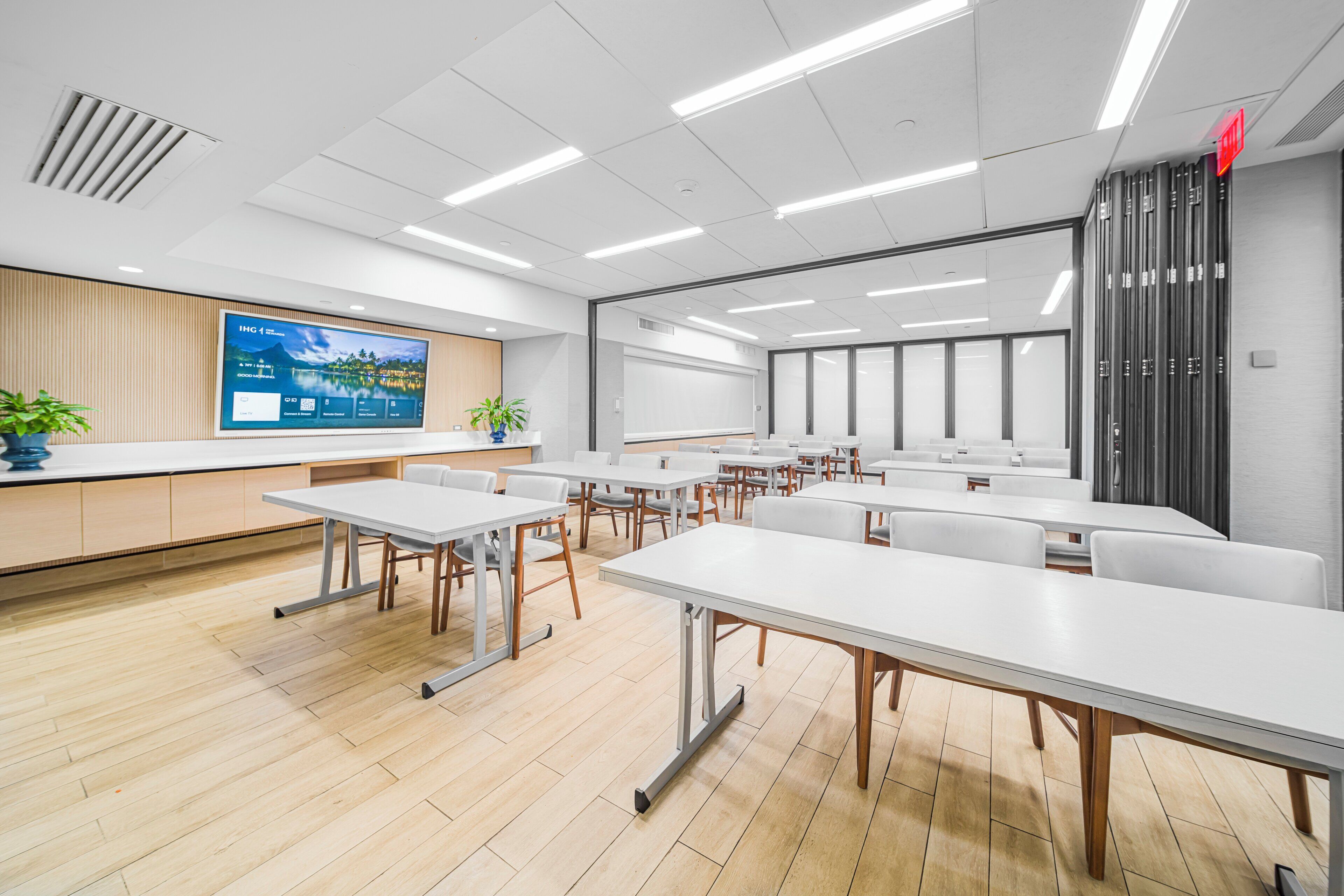 Una sala de conferencias moderna con mesas y sillas blancas, una pantalla grande y un panel de pared de madera.