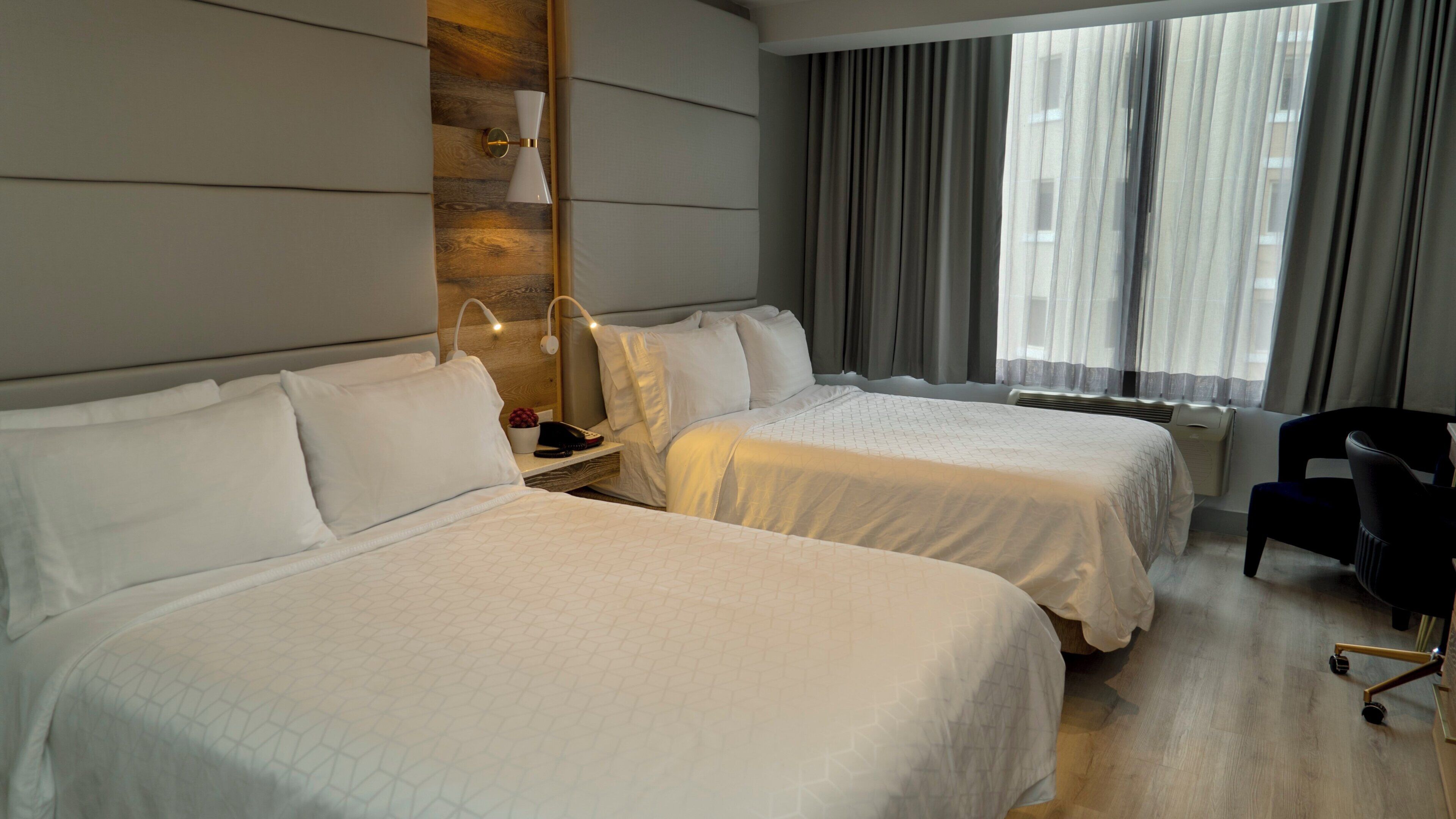 A hotel room with two beds, a desk with a lamp, a chair, and a window with curtains.