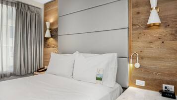 Una habitación de hotel moderna con una cama grande, un cabecero de madera y una mesita de noche con una lámpara y un reloj.