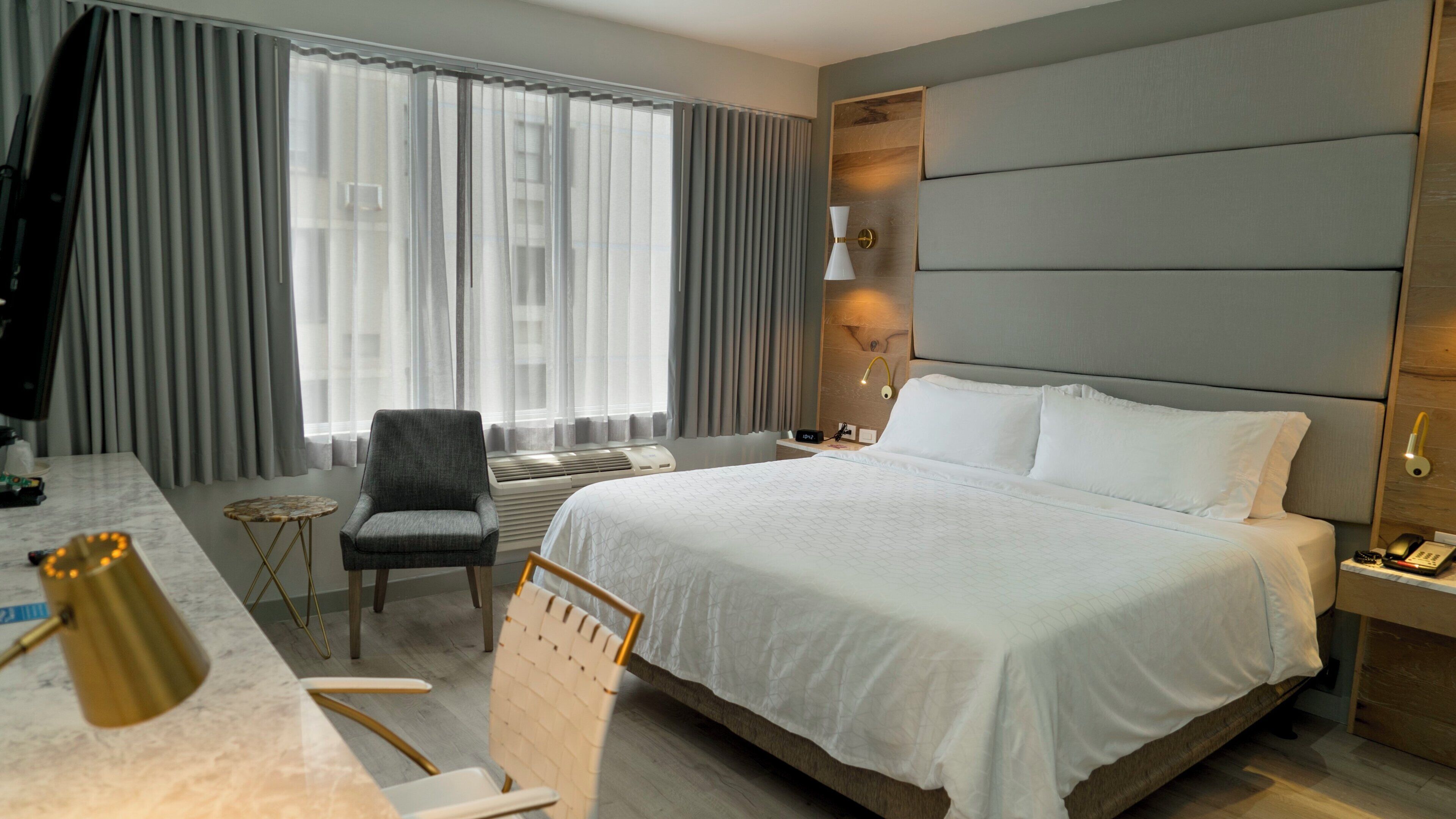 A hotel room with a large bed, a desk with a lamp, a chair, and a small table.