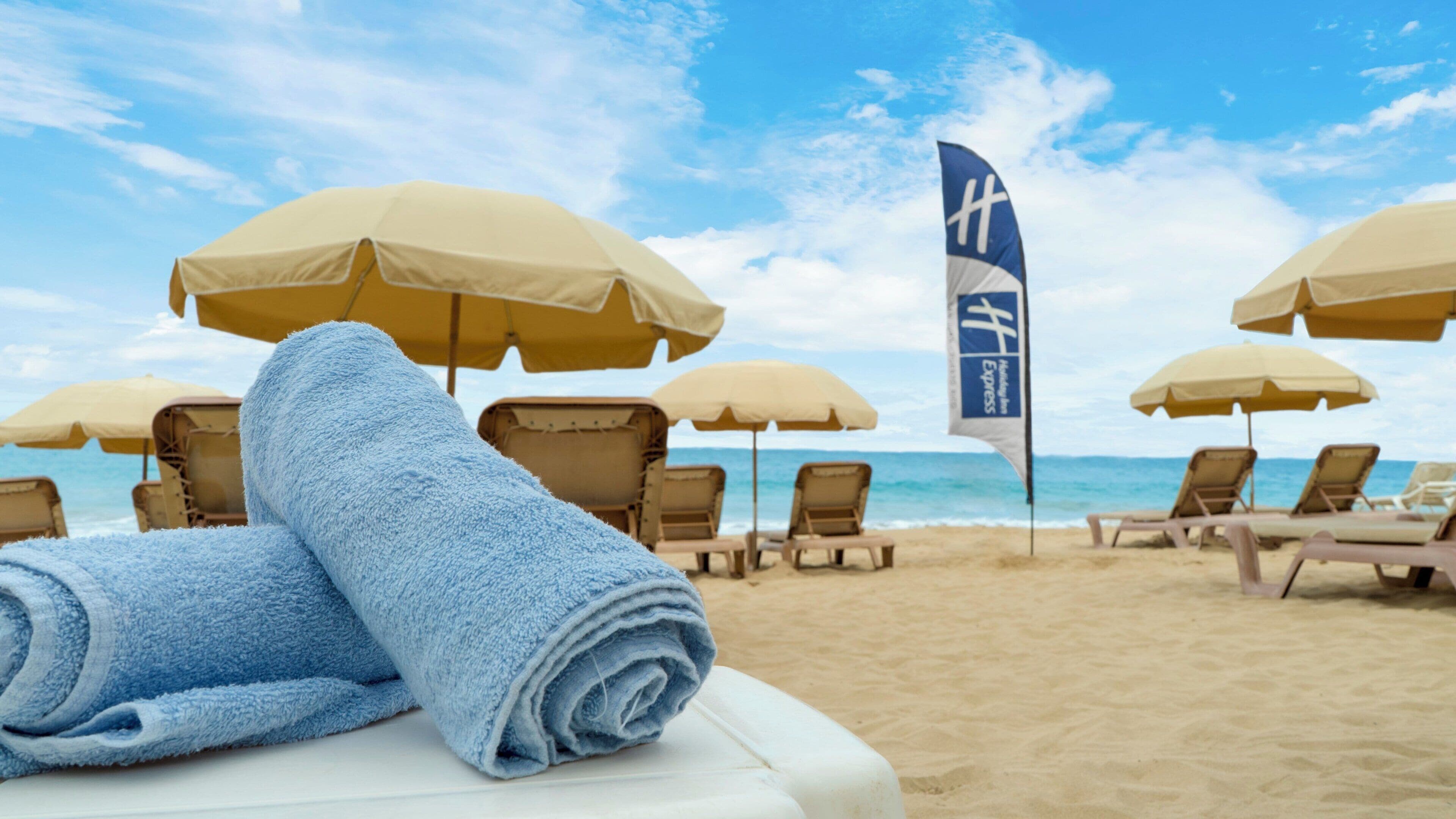 A beach setting with sun loungers and umbrellas, a flag, and a rolled towel on a white surface.