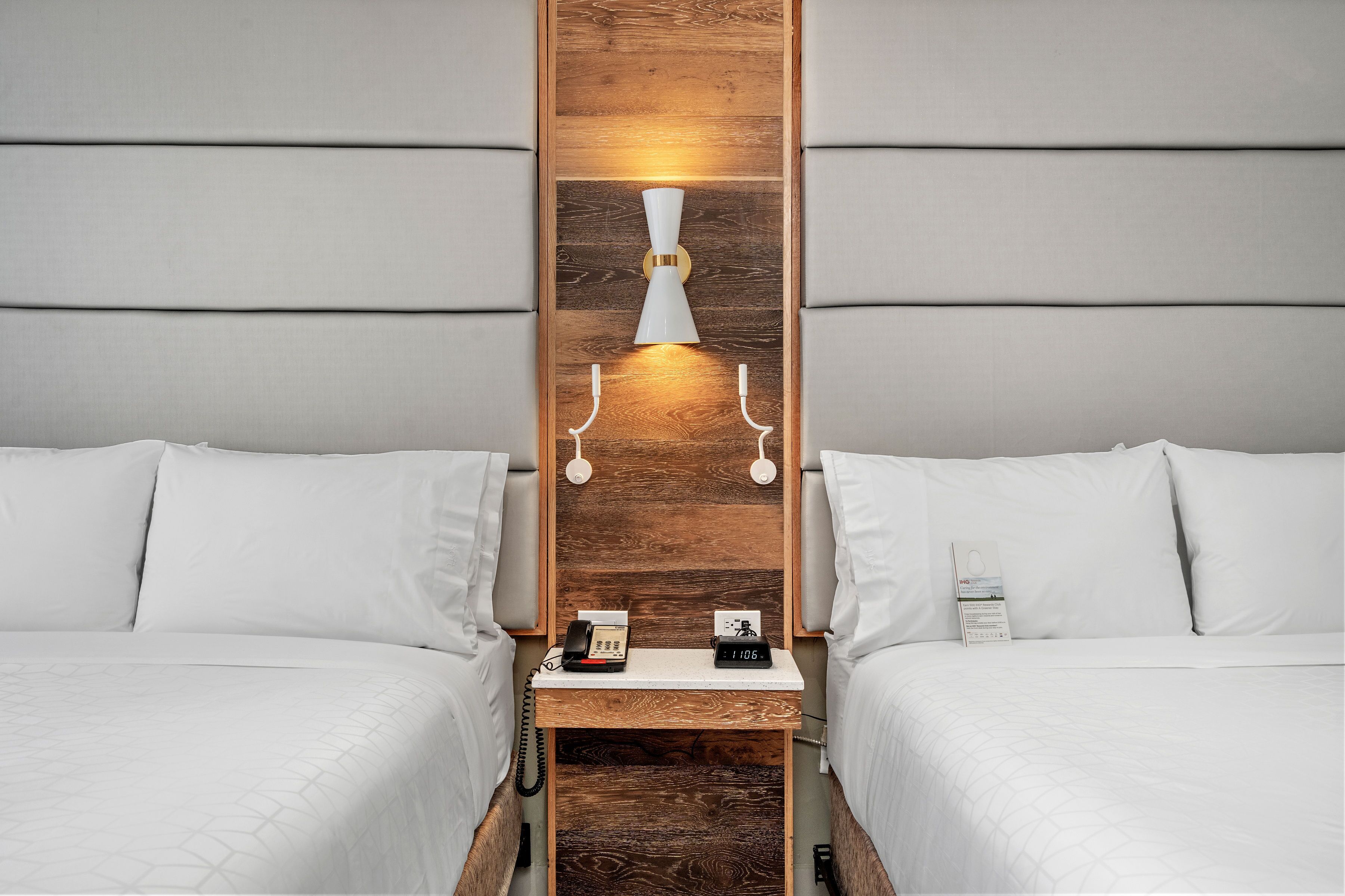 A hotel room with two beds, a wooden headboard, a bedside table with a phone and a clock, and a wall-mounted light fixture.