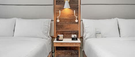 A hotel room with two beds, a wooden headboard, a bedside table with a phone and a clock, and a wall-mounted light fixture.