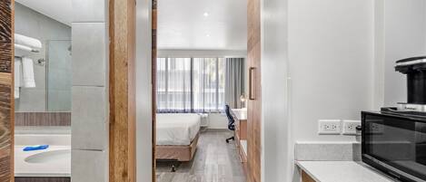 A modern hotel room with a wooden floor, a bed, a bathroom with a bathtub, and a kitchenette with a microwave.