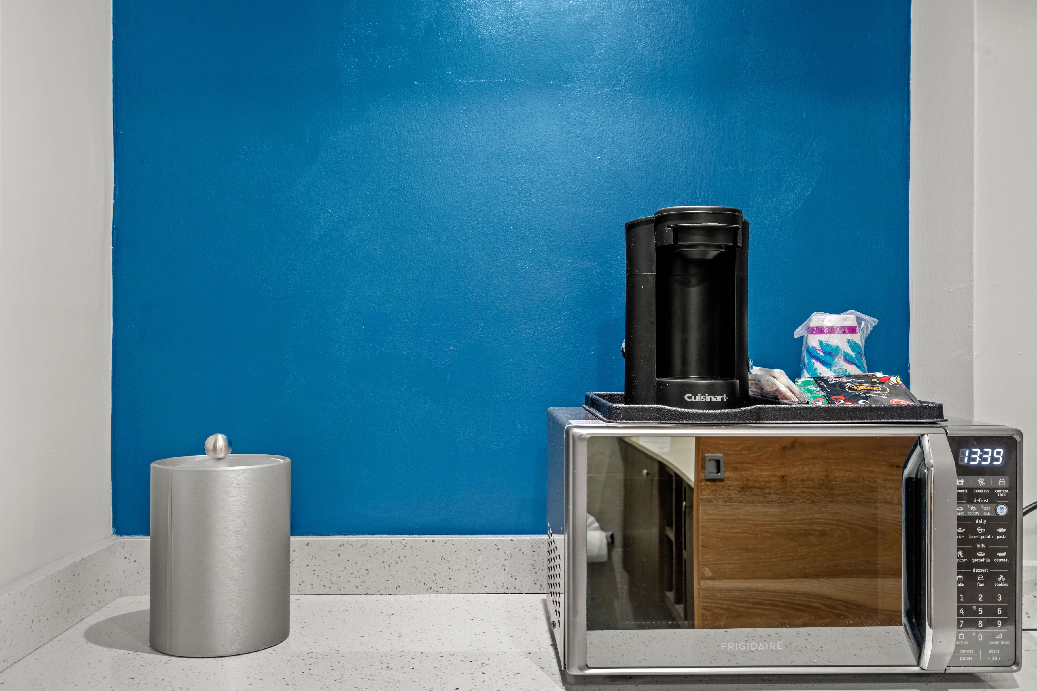 Un four à micro-ondes avec un panneau décoratif en bois, une machine à café et une poubelle, le tout posé sur un plan de travail devant un mur bleu.