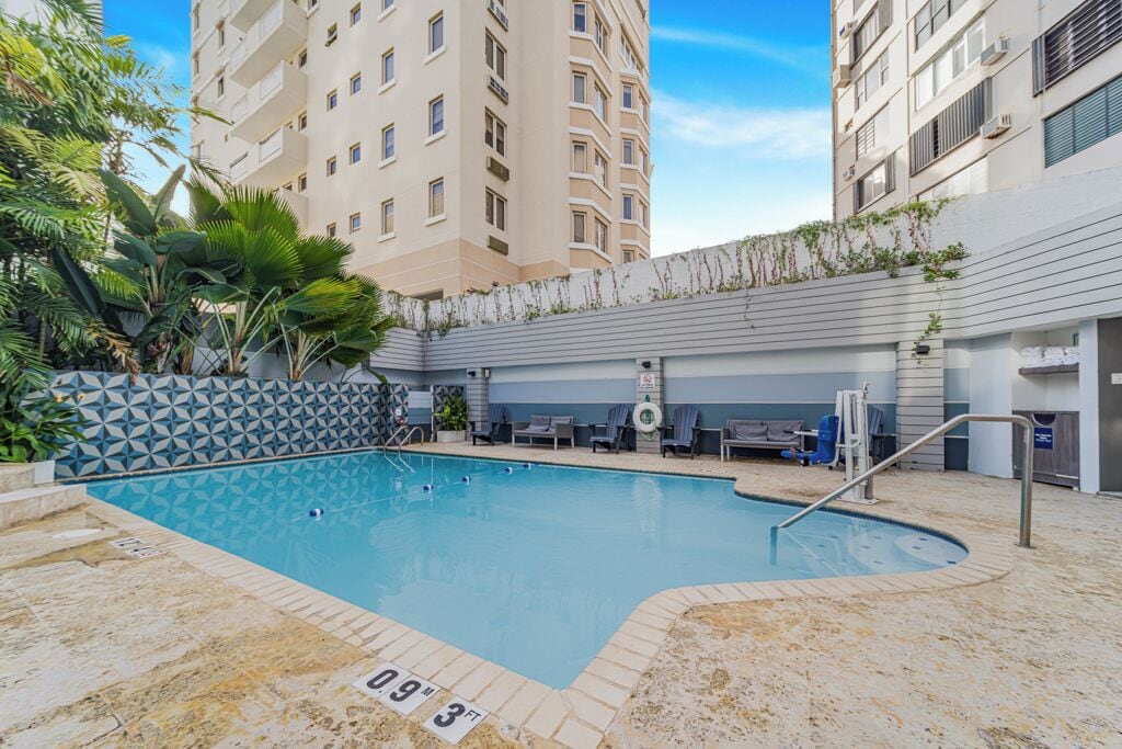 A swimming pool with a geometric tile pattern, surrounded by lounge chairs and a low wall with climbing plants.