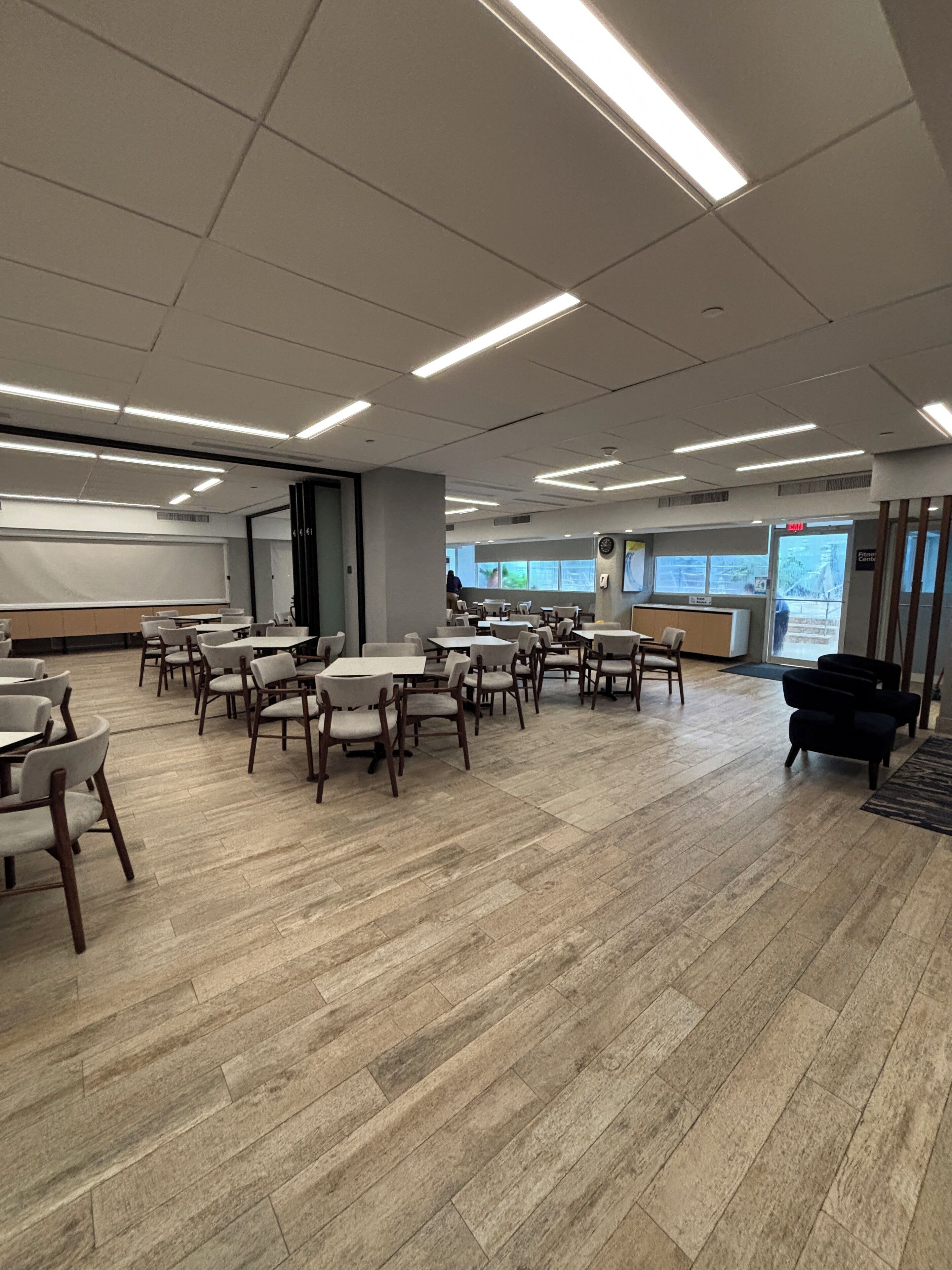 A spacious room with multiple tables and chairs, a whiteboard, and a view of the outdoors through large windows.