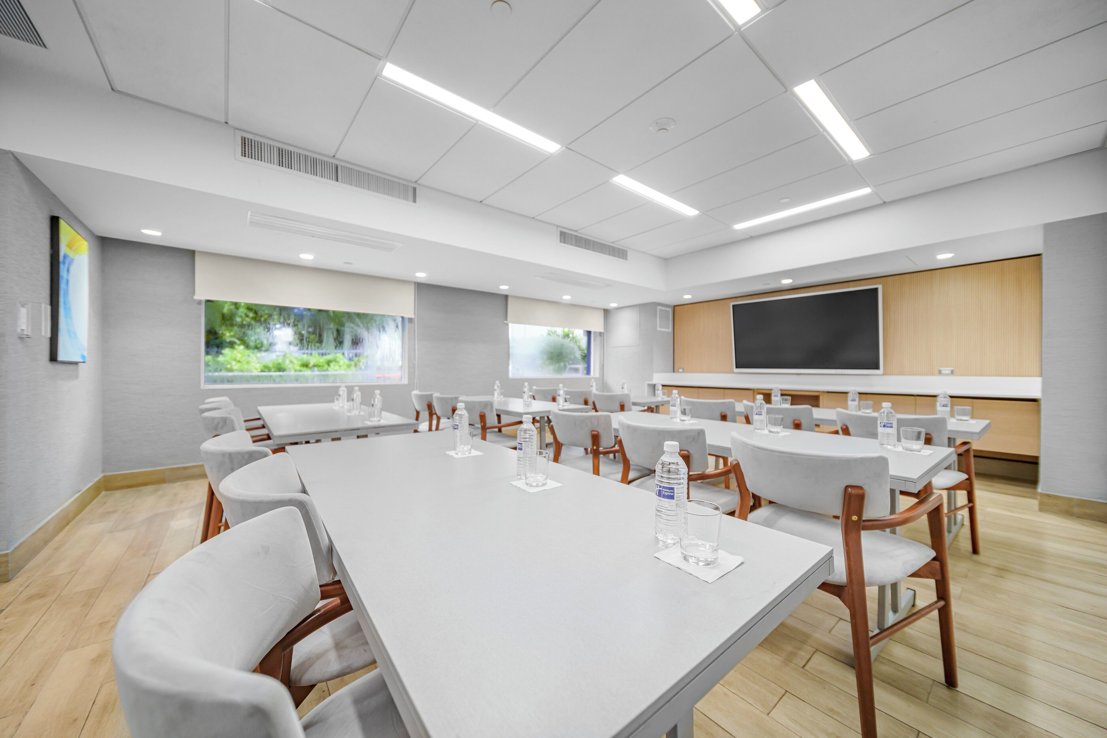 Una sala de conferències amb taules i cadires blanques, una pantalla gran i finestres amb vista a vegetació.