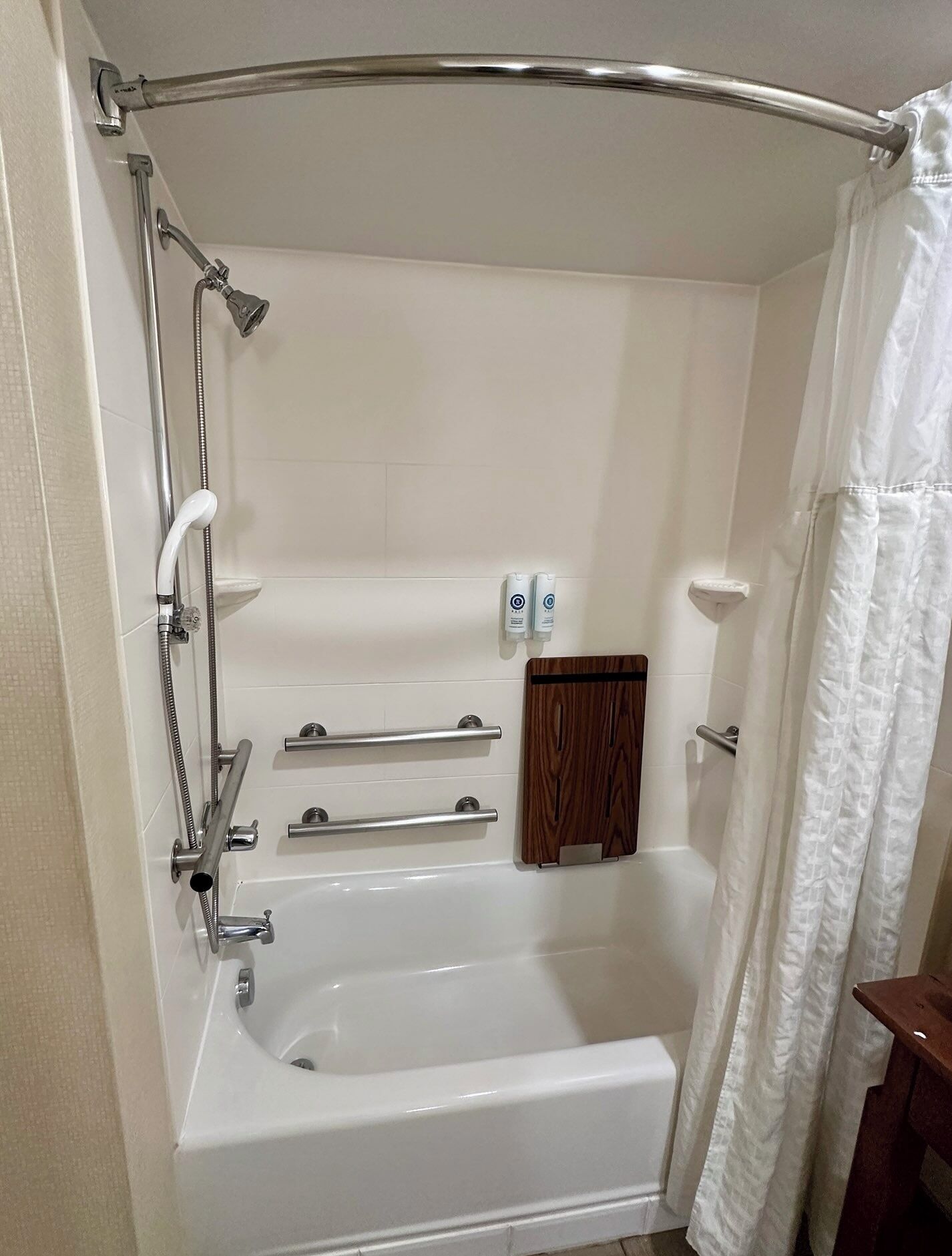 A bathroom with a bathtub, shower, grab bars, and a wooden shelf.