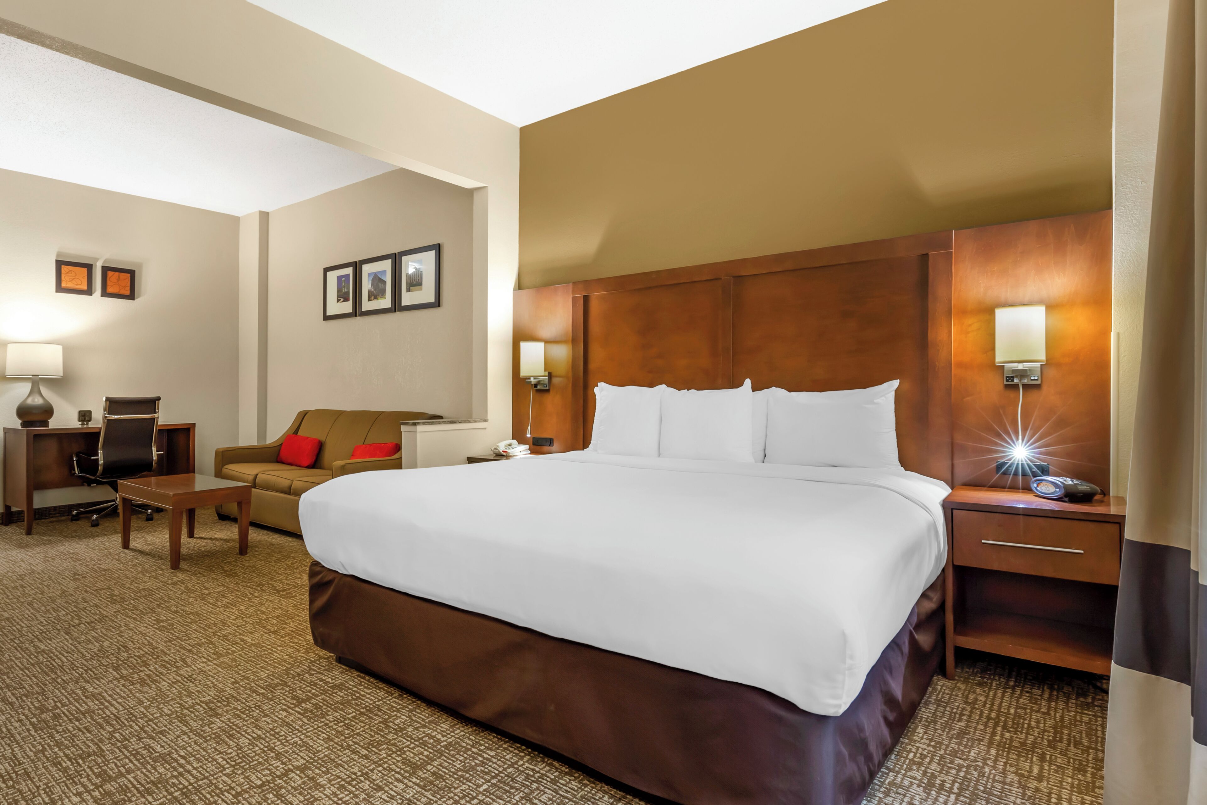 A hotel room with a large bed, a desk with a chair, a sofa, and a nightstand with a lamp.