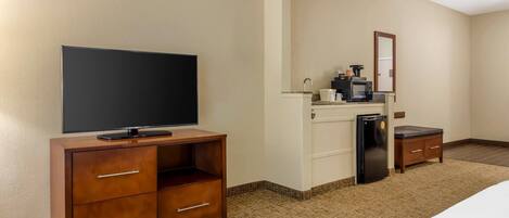 A hotel room with a flat-screen TV on a wooden cabinet, a kitchenette with a microwave and coffee maker, a refrigerator, and a small table.