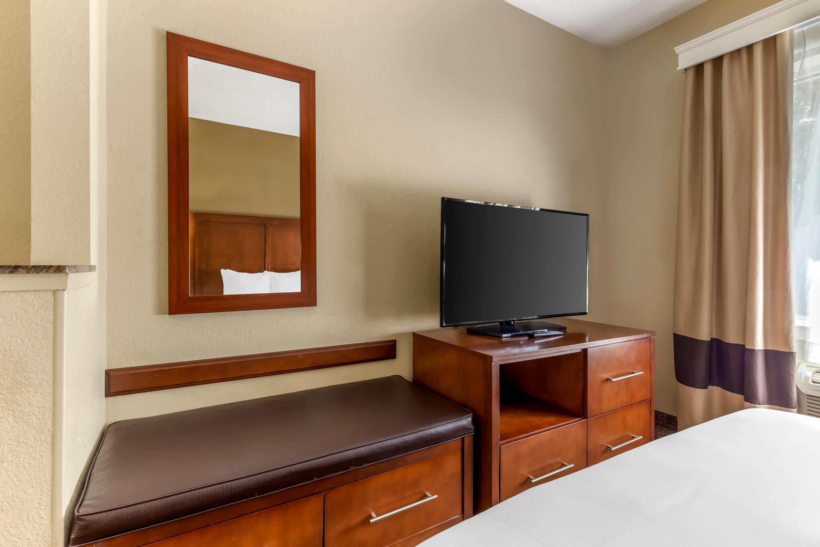 A hotel room with a bed, a wooden dresser, a television, a bench, and a mirror.