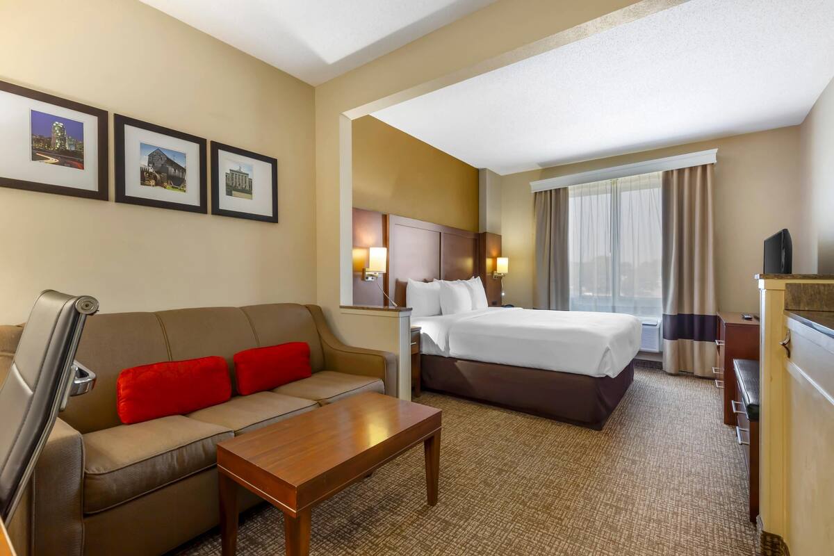 A hotel room with a bed, a sofa, a coffee table, and a desk. There are framed pictures on the wall and a window with curtains.