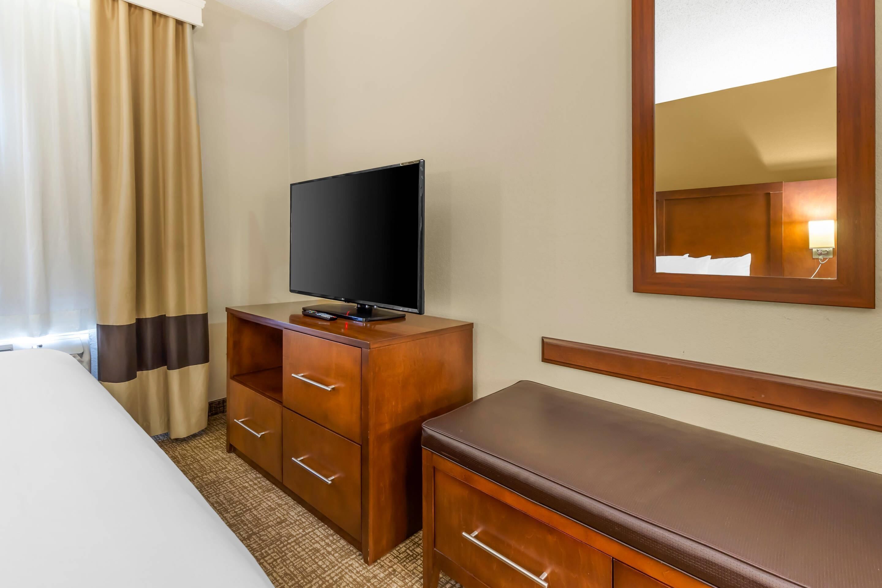 A hotel room with a bed, a wooden dresser with a flat-screen TV, a mirror, and a window with curtains.