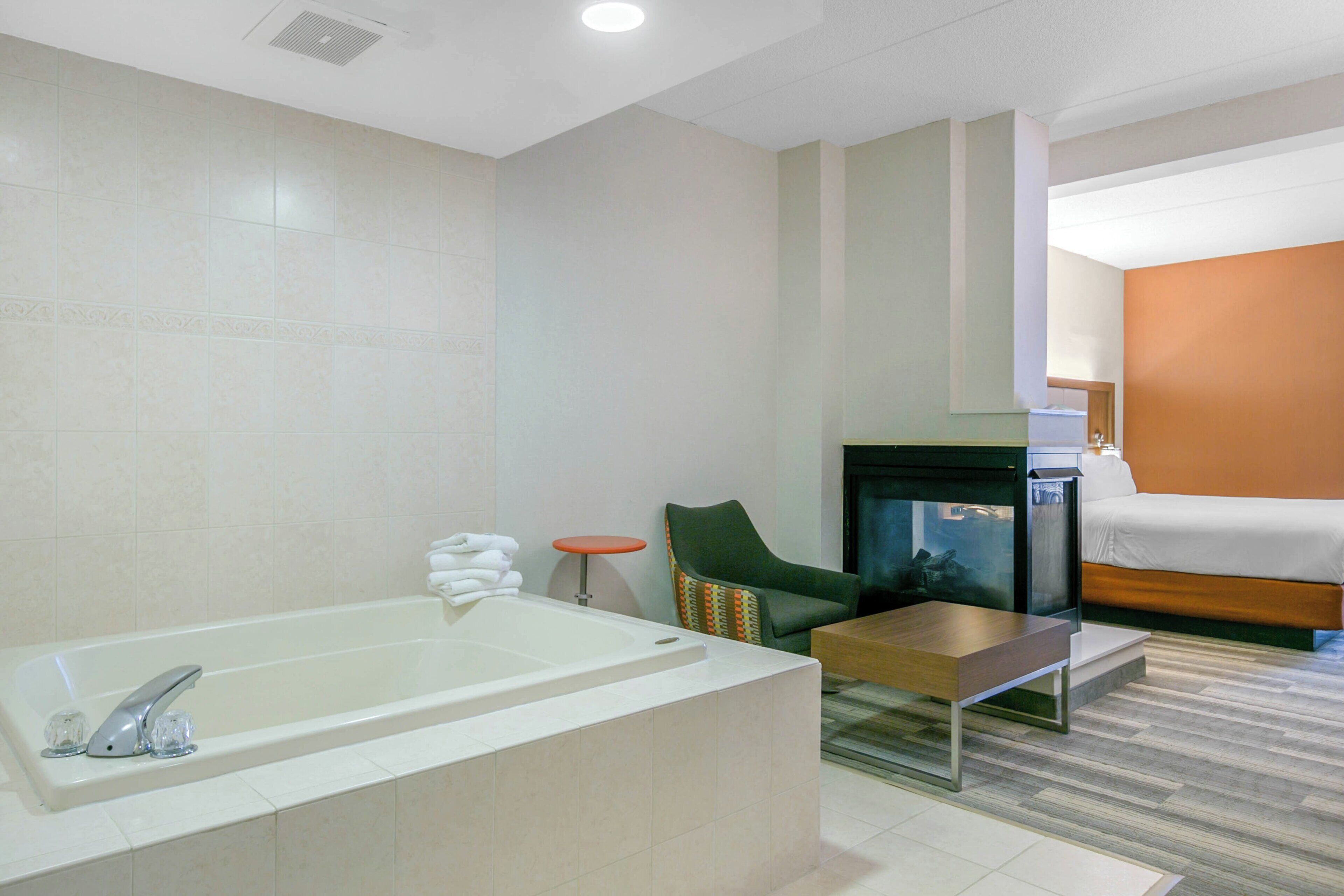 A modern hotel room with a bathtub, a small table, a chair, a sofa, and a fireplace.