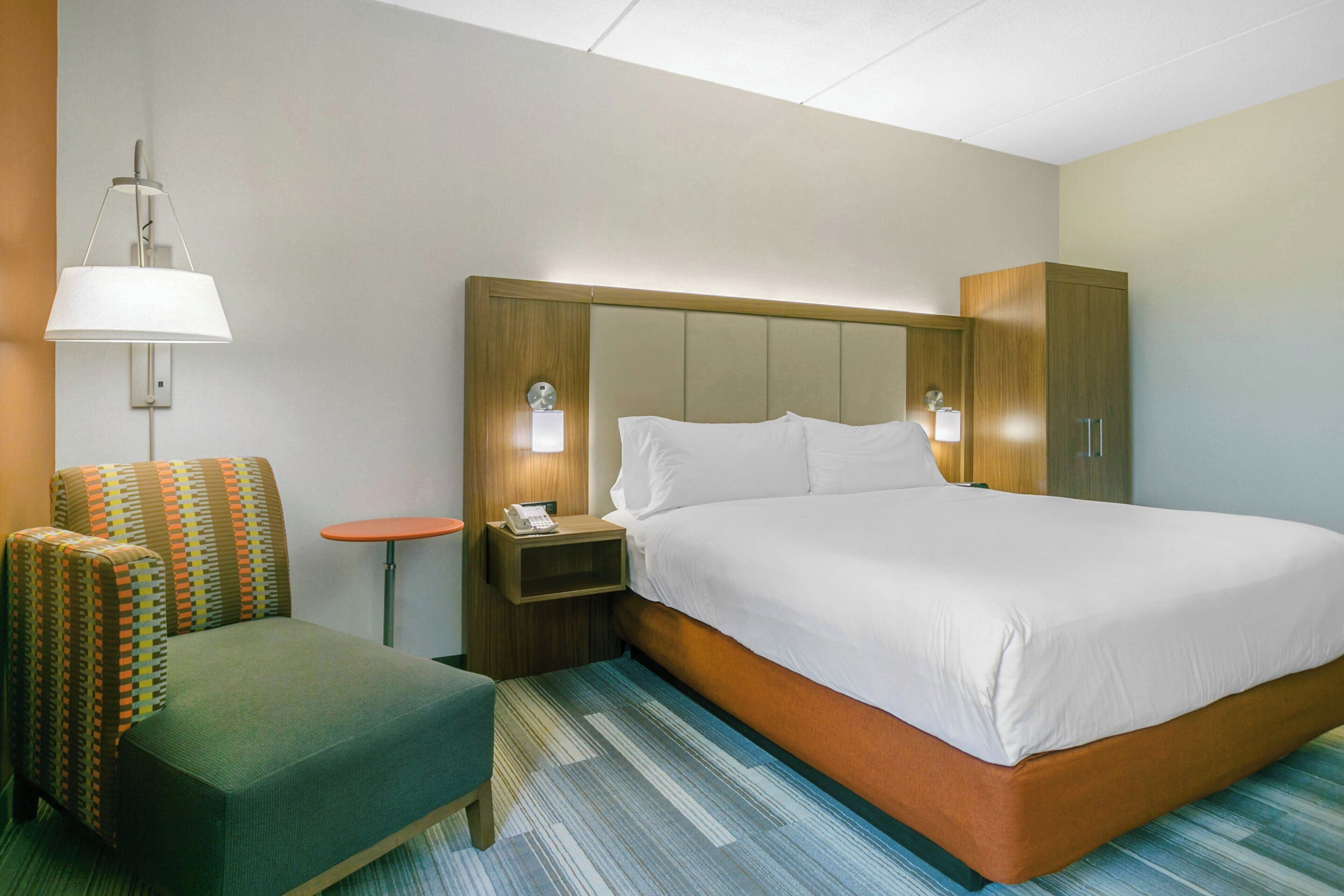 A hotel room with a bed, a nightstand, a lamp, a chair, and a wardrobe.