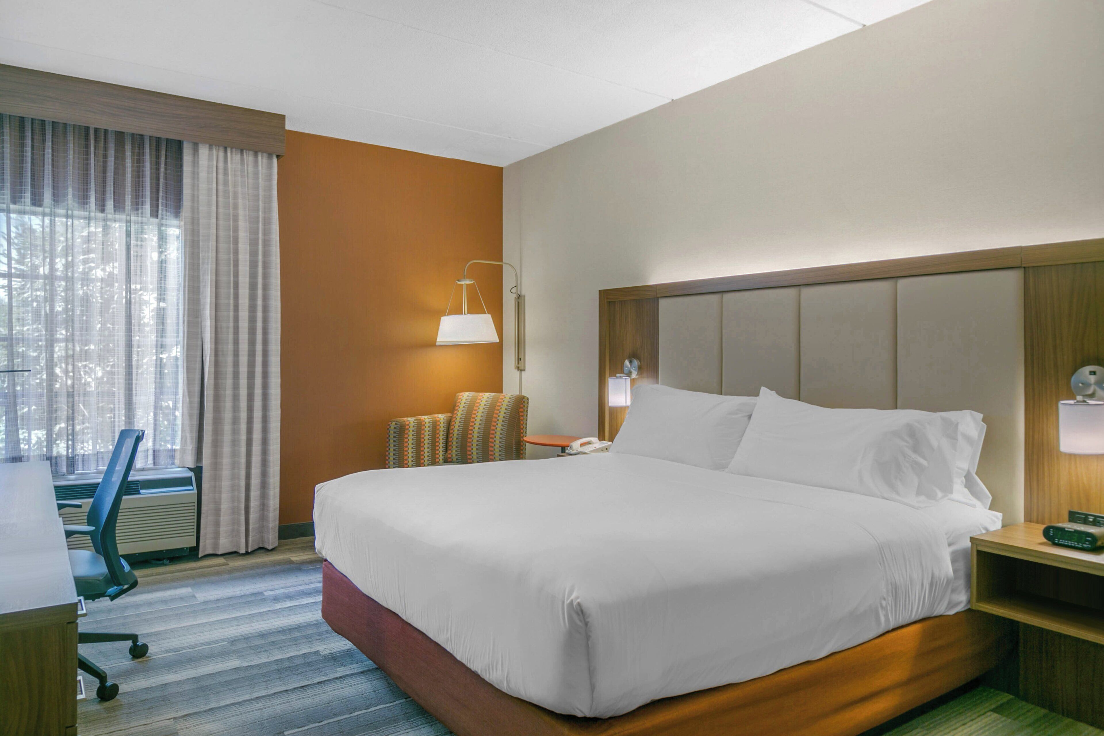 A hotel room with a large bed, a desk, a chair, a lamp, and a window with curtains.