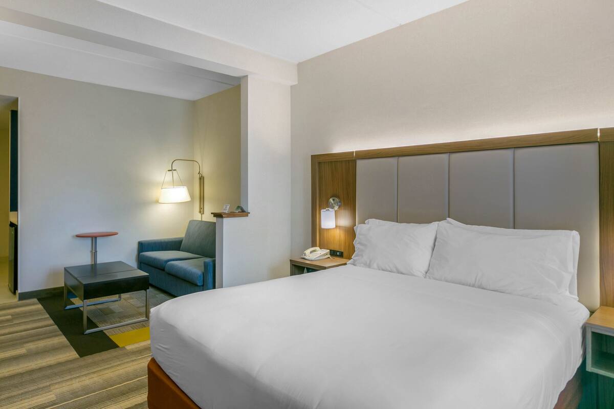A hotel room with a large bed, a desk, a chair, and a nightstand with a lamp and phone.