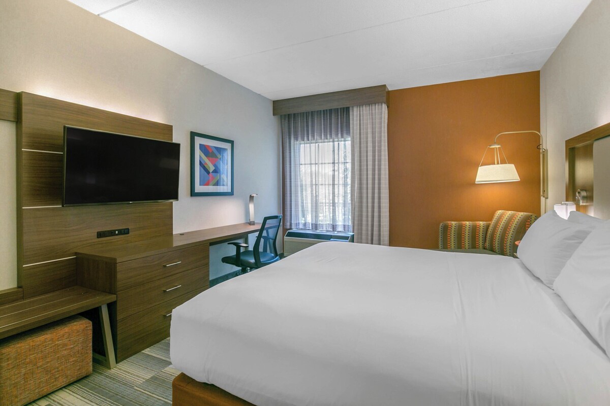A hotel room with a bed, a desk, a chair, a TV, a window with curtains, and a lamp.