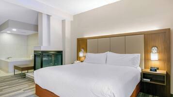 Habitación de hotel con una cama grande, una mesita de noche con lámpara, un tocador con reloj, un baño con lavamanos y espejo, y una pequeña zona de estar.