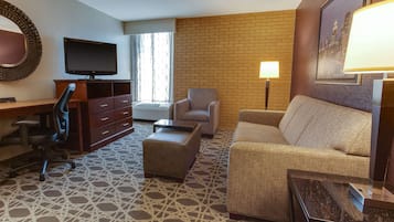 A hotel room with a flat-screen TV, a sofa, a chair, a desk with drawers, a patterned carpet, and a brick wall.