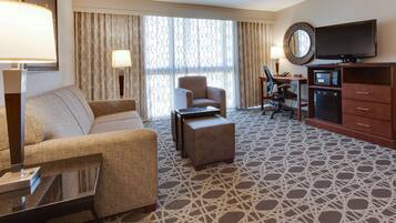 A hotel room with a sofa, armchair, ottoman, TV, microwave, and a desk with a lamp.
