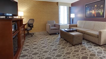 Habitación de hotel con pared de ladrillo, un televisor de pantalla plana grande, un sofá, una silla, una mesa de centro y una fotografía enmarcada de un paisaje urbano.
