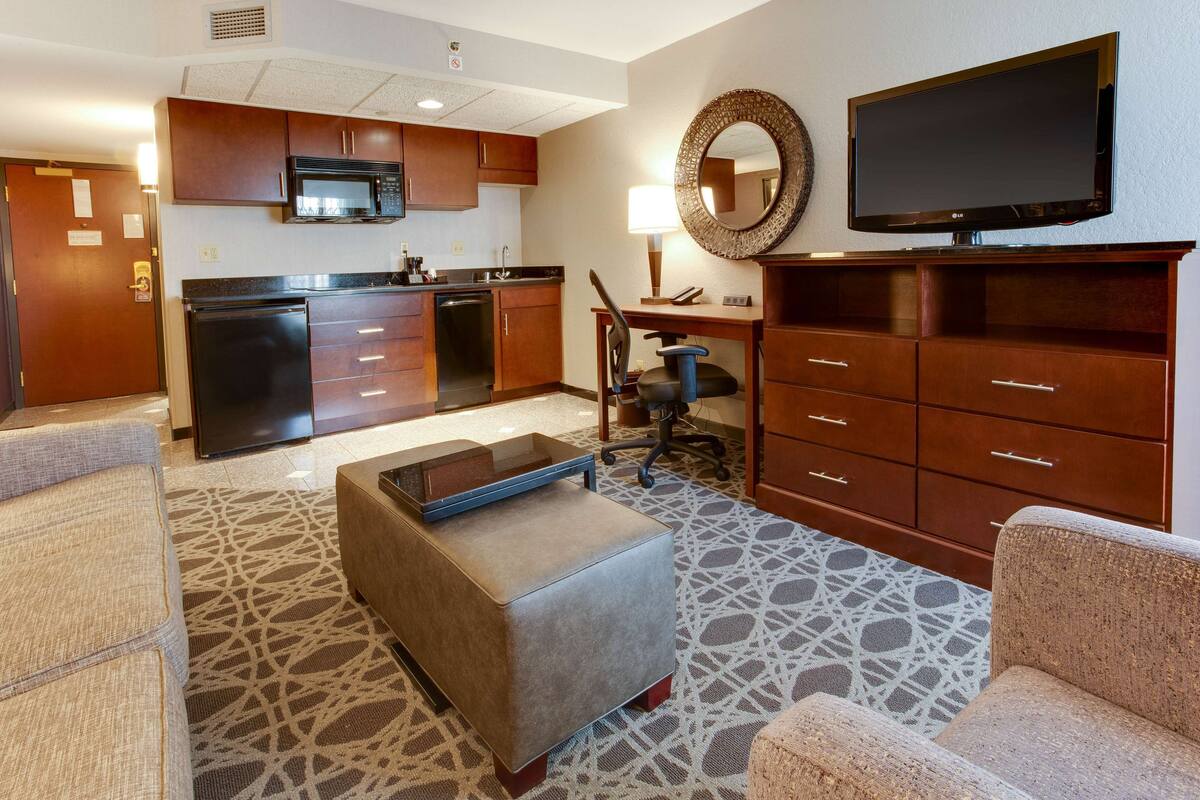 Habitación de hotel con kitchenette, televisión de pantalla plana, escritorio con silla y alfombra estampada.