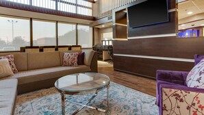 Amplia sala de estar con un sofá, una mesa de centro de vidrio y un sillón morado. Un televisor de pantalla plana está montado en la pared, y se observa un escritorio con una computadora al fondo.