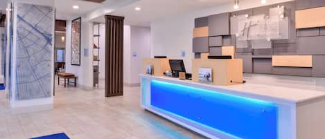 A hotel reception area with a modern design, featuring a reception desk with a blue illuminated panel, a map on the wall, and a seating area with a bench.