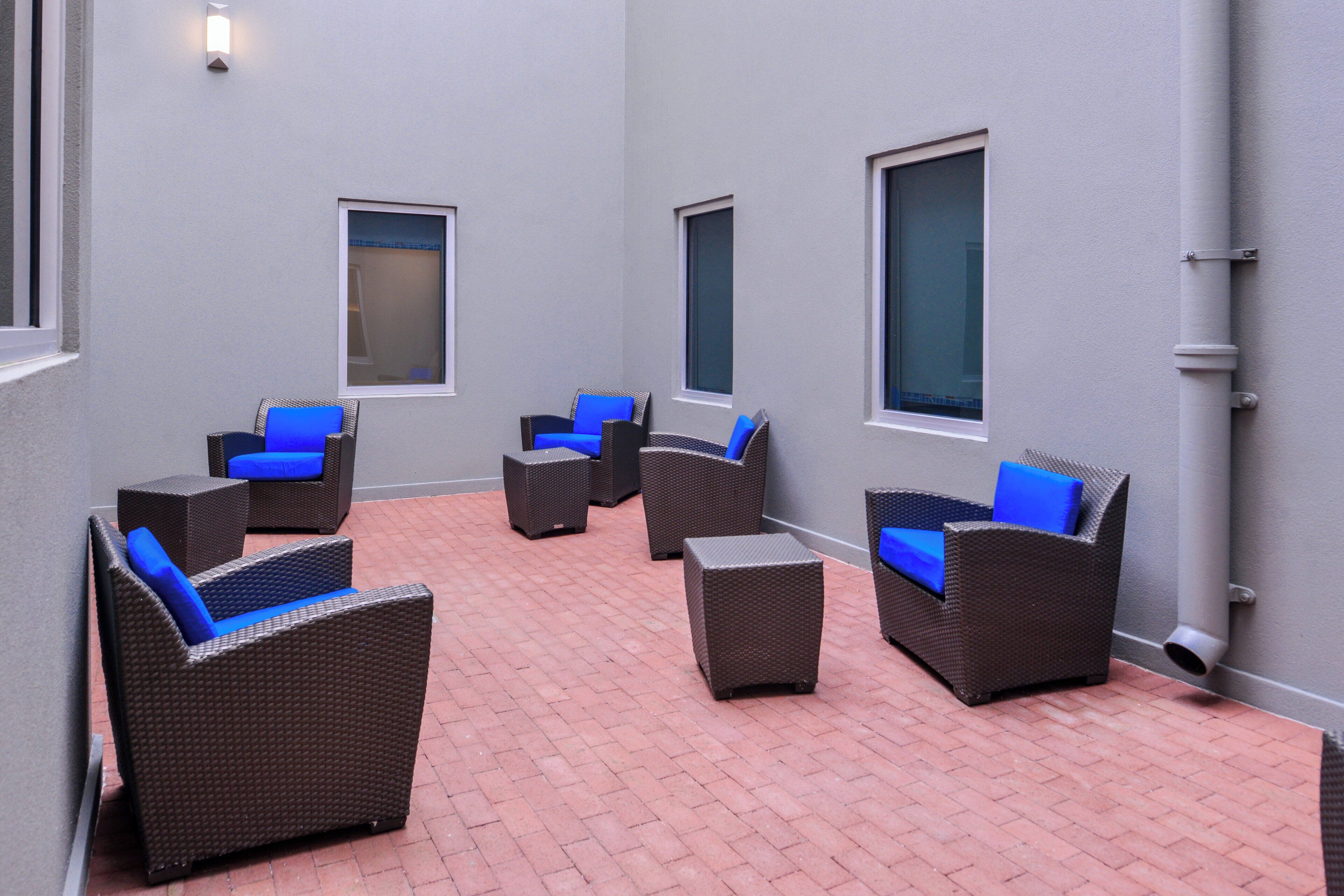A patio area with wicker furniture and blue cushions, a tiled floor, and a grey wall with windows.