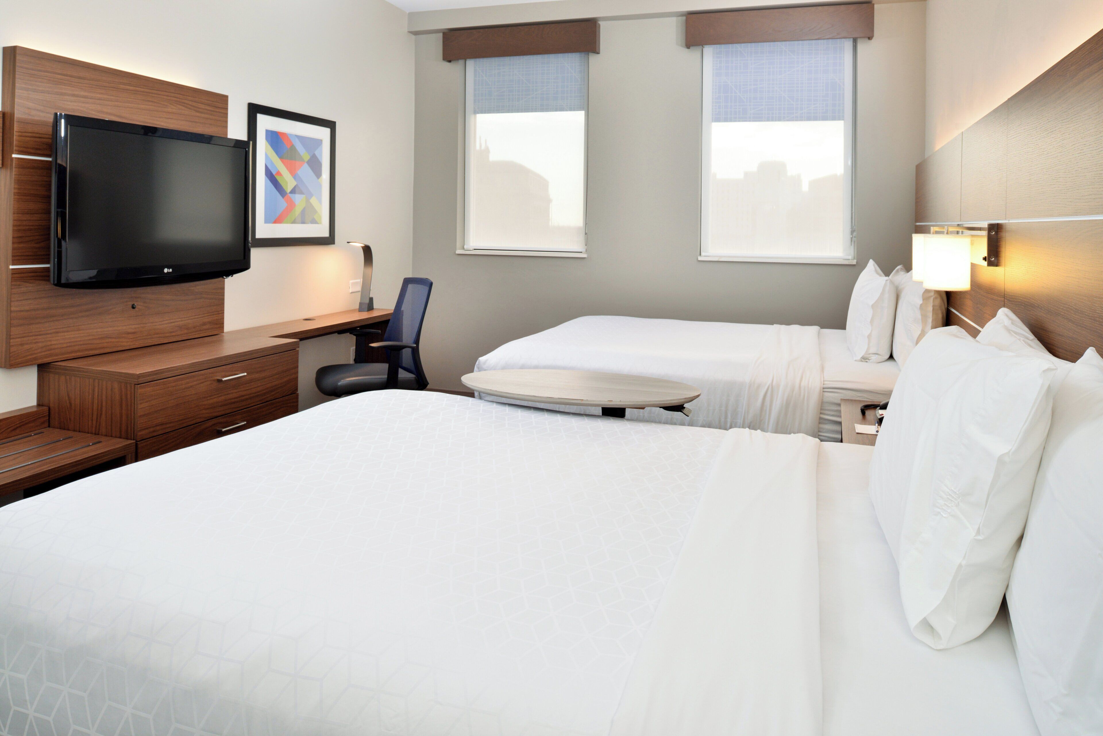 A hotel room with a bed, a desk, a chair, a TV, and two windows.