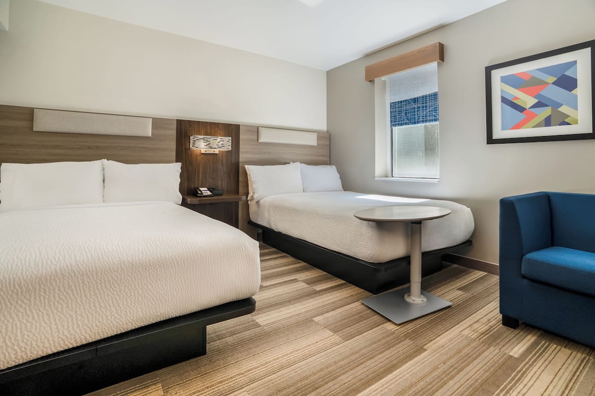 A hotel room with two beds, a small table, a blue armchair, a nightstand, and a window with blinds.