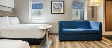 Una habitación de hotel con una cama, un sofá azul, una mesita redonda y dos ventanas con persianas.