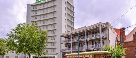 A multi-story hotel with a balcony on the second floor, a trolley in front, and a tree in the foreground.