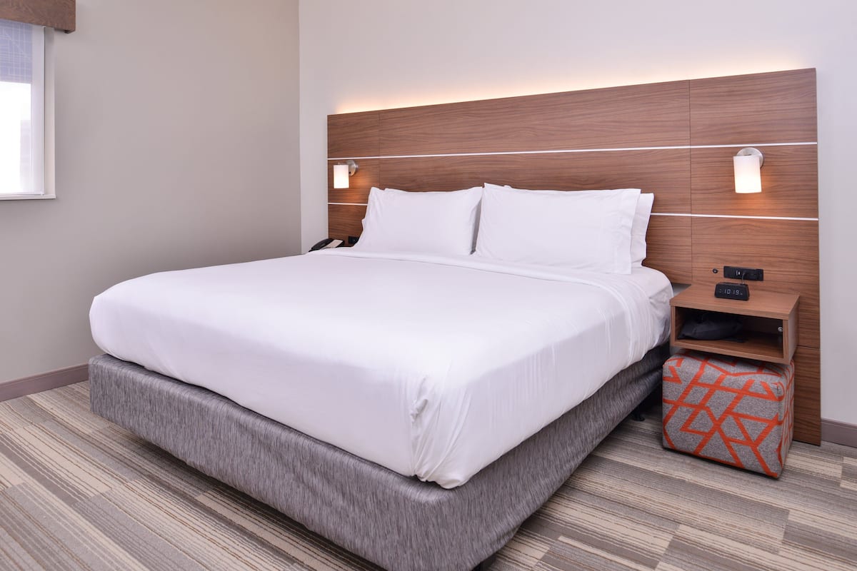 A neatly made bed with a wooden headboard and bedside lamps, a small wooden nightstand, and a patterned ottoman.
