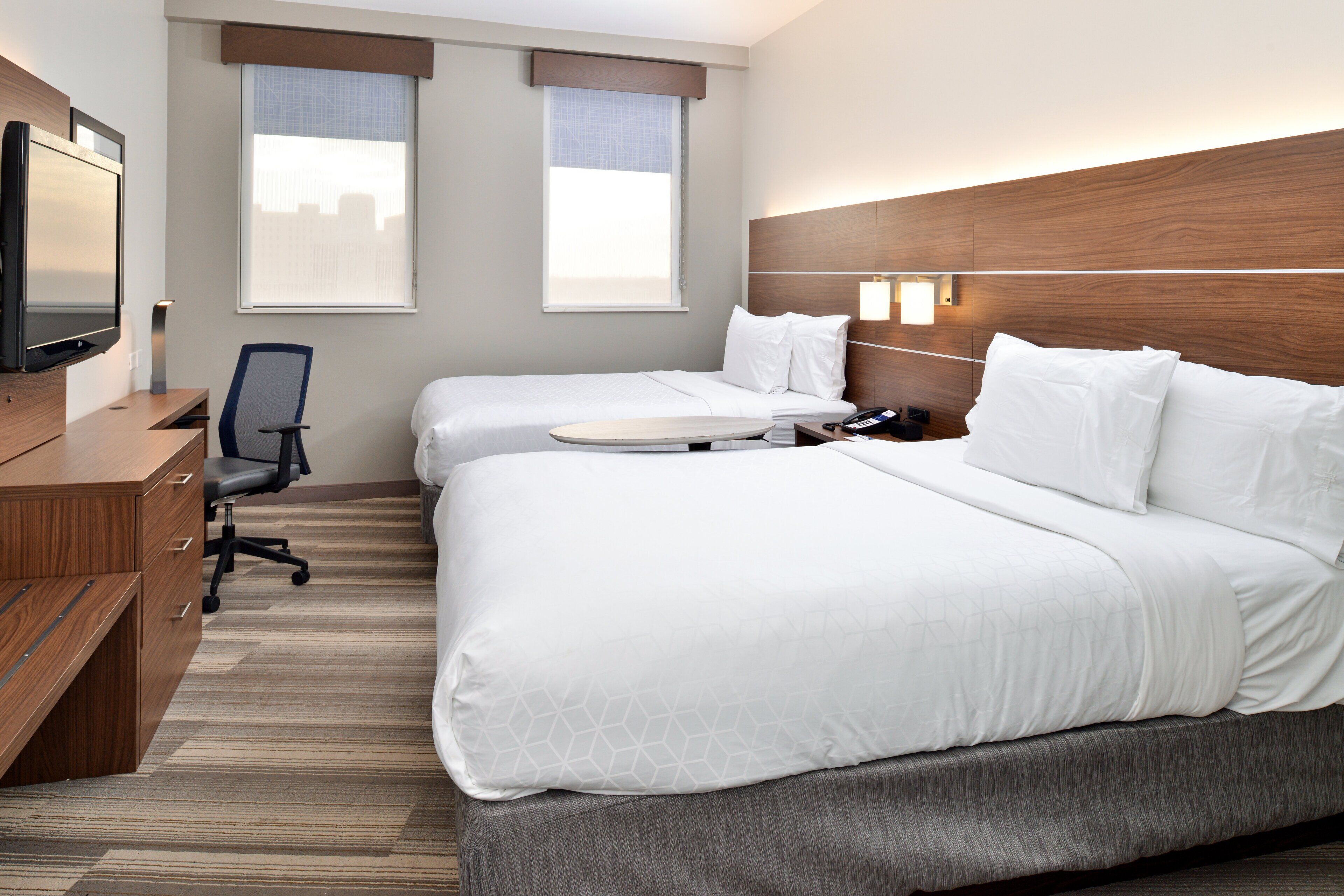 A hotel room with two beds, a desk with a chair, a TV, and a window with blinds.