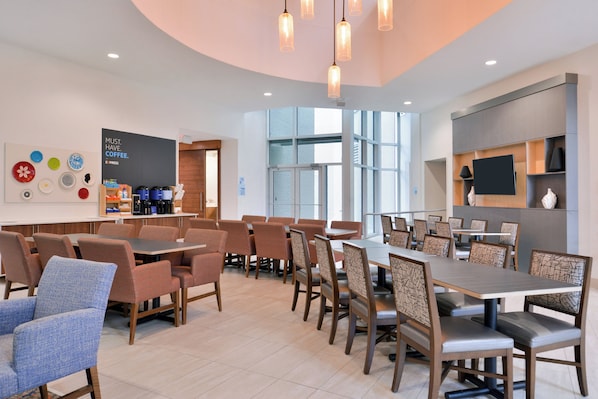 A modern dining area with multiple tables and chairs, a coffee station, and large windows providing natural light.