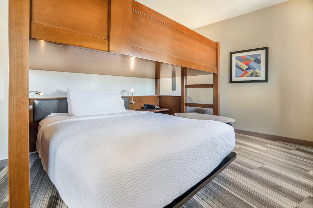 A hotel room with a large bed, wooden headboard, bedside tables, a lamp, and a framed abstract art piece on the wall.