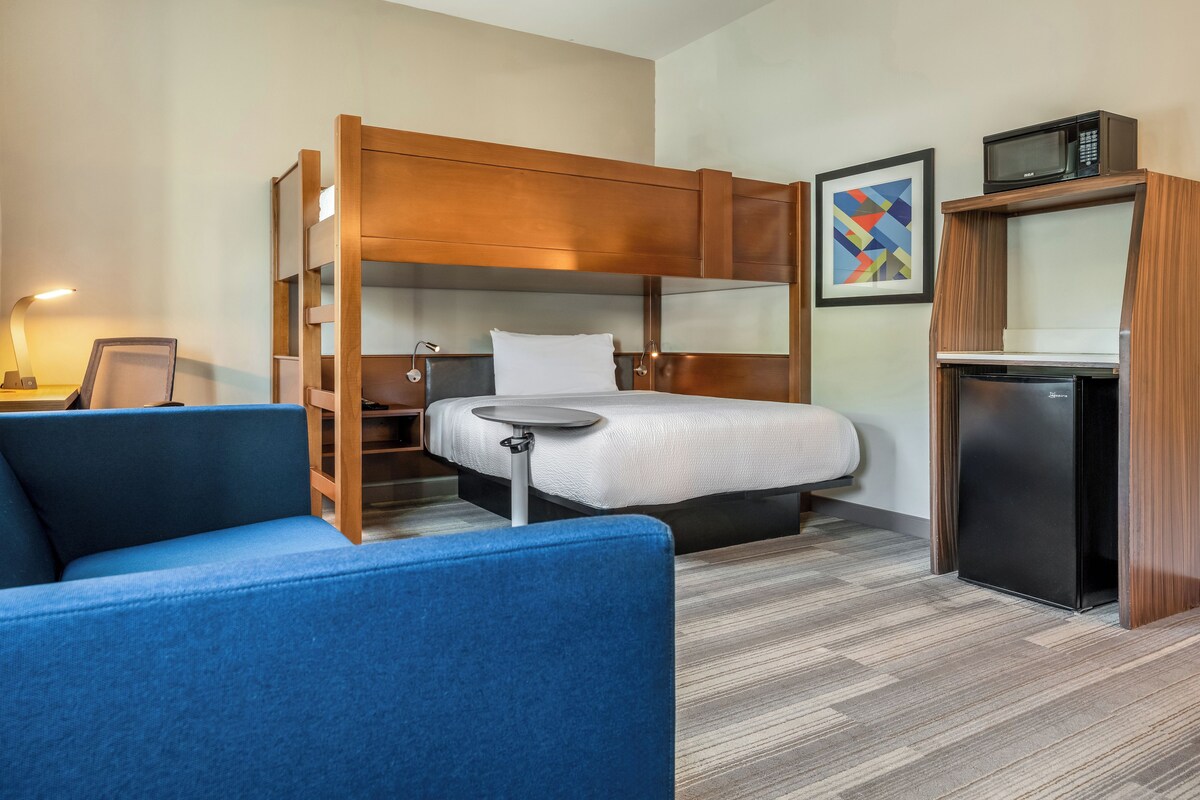 A compact hotel room with a bunk bed, a small desk, a chair, a microwave, and a refrigerator.