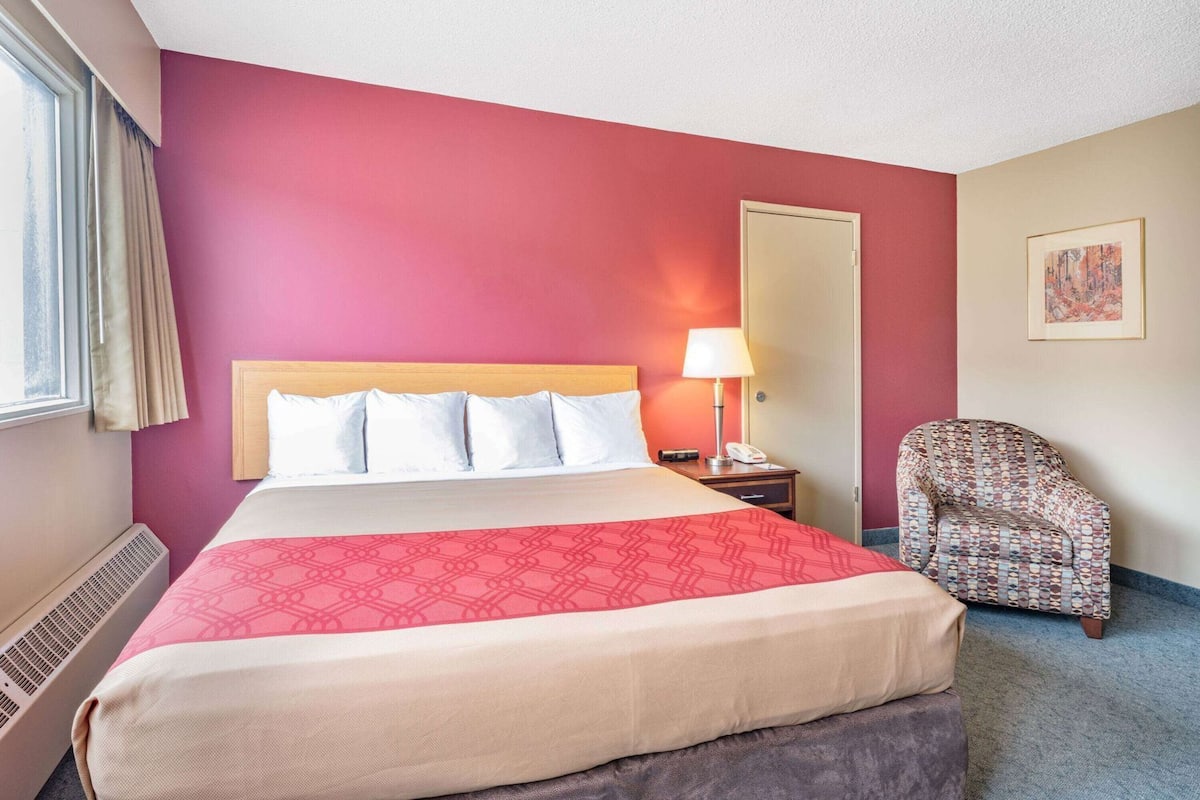 Una habitación de hotel con cama, mesita de noche, silla, puerta y ventana con cortinas.
